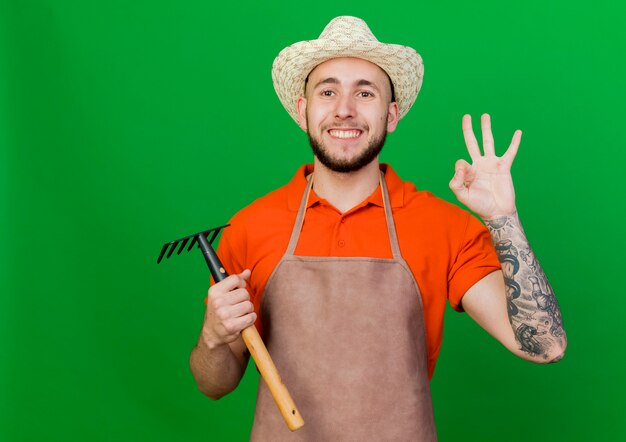 Glimlachende tuinman man met tuinhoed gebaren ok hand teken en houdt hark