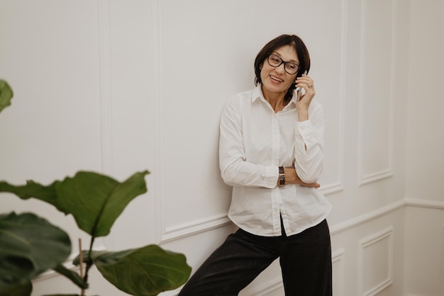 Glimlachende volwassen blanke brunette vrouw in wit overhemd en zwarte broek gebruikt haar telefoon Gesprek concept