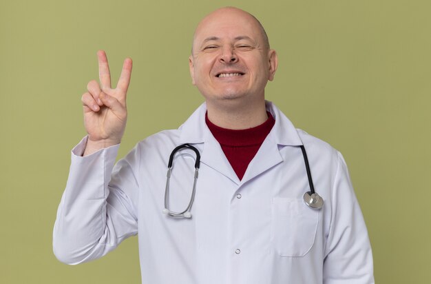 Glimlachende volwassen man in doktersuniform met stethoscoop gebaren overwinningsteken