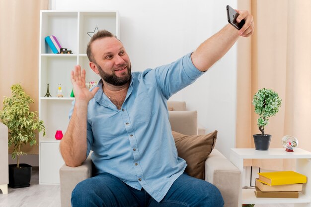 Glimlachende volwassen Slavische man zit op fauteuil hand opheffen en kijken naar telefoon in de woonkamer