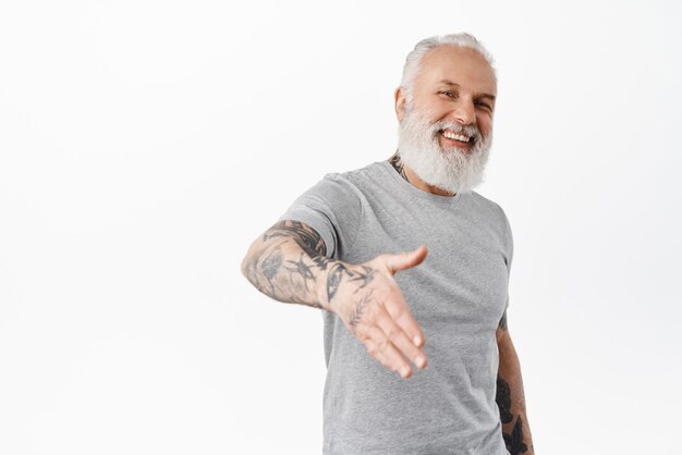 Glimlachende vriendelijke volwassen man met tatoeages geeft hand voor handdruk en lacht leuk om je te ontmoeten gebaar iemand te ontmoeten die je begroet terwijl hij op een witte achtergrond staat