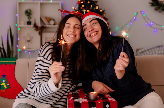 Glimlachende vrij jonge meisjes met rendierglazen en kerstmuts met wonderkaarsen zittend op fauteuils en genieten van kersttijd thuis