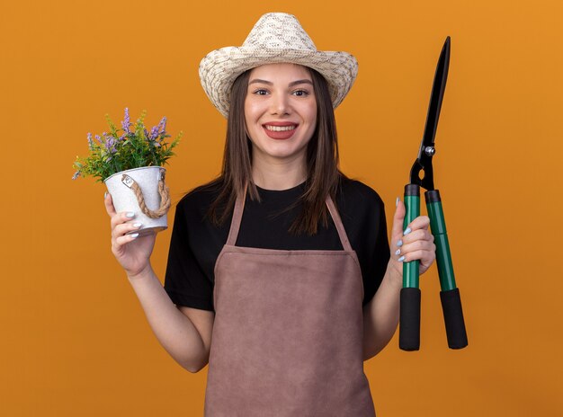 Glimlachende vrij kaukasische vrouwelijke tuinman die het tuinieren hoed draagt ?? die bloempot houdt en tuinierende schaar die op oranje muur met exemplaarruimte wordt geïsoleerd