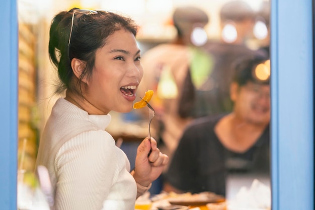 Glimlachende vrolijke Aziatische vrouwelijke hand houdt fish and chips-ontbijt terwijl ze geniet van het eten van een lachgesprek met haar vriend in de buurt van de raamwinkel in café-restaurant