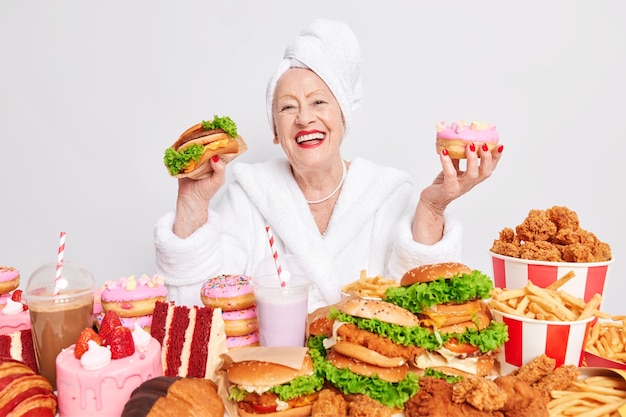 Glimlachende vrolijke oude vrouw voelt zich erg gelukkig houdt heerlijke hamburger en donutdragers badjas en handdoek op hoofd eet junkfood