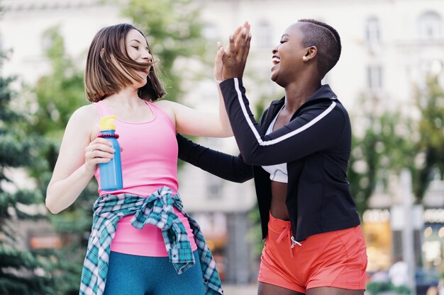 Glimlachende vrolijke vrienden in sportkleding die in de stad rennen en discussiëren over het geven van hallo vijf vieren van succes en geluk Multi-etnische vrouwen met een fitnesstraining