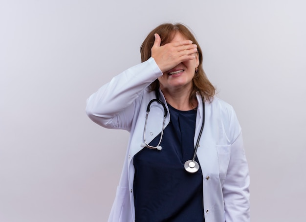 Glimlachende vrouw arts die op middelbare leeftijd medische mantel en stethoscoop sluitende ogen met hand op geïsoleerde witte muur met exemplaarruimte draagt