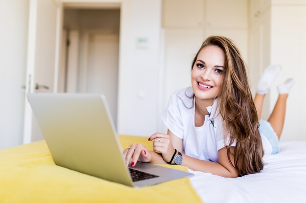 Glimlachende vrouw die haar sociale media inhaalt terwijl ze in bed ontspant met een laptop op een luie dag thuis