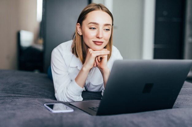 Glimlachende vrouw die haar sociale media inhaalt terwijl ze op een luie dag in bed ontspant met een laptopcomputer