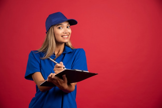 Glimlachende vrouw die in blauw uniform met potlood op klembord schrijft.