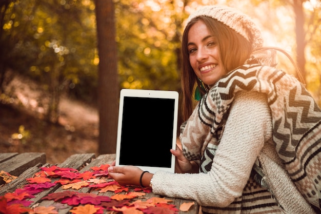 Gratis foto glimlachende vrouw die lege tablet in de herfstpark toont
