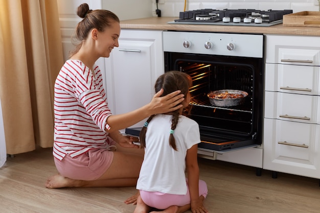 Glimlachende vrouw met haarverbod die het hoofd van haar dochtertje aanraakt terwijl het kind achteruit naar de camera zit en naar de oven kijkt met bakken, het vrouwtje kijkt naar het kind met liefde, samen koken.