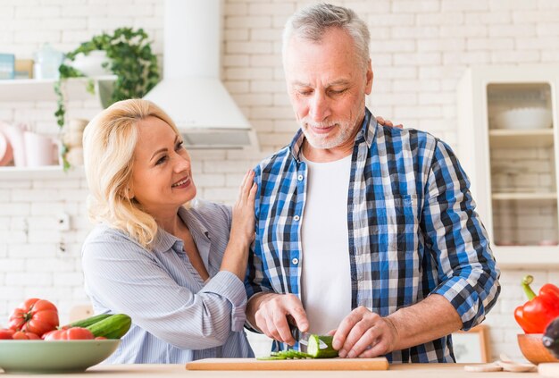 Glimlachende vrouw ondersteunend haar echtgenoot die de komkommer met mes op lijst in de keuken snijdt