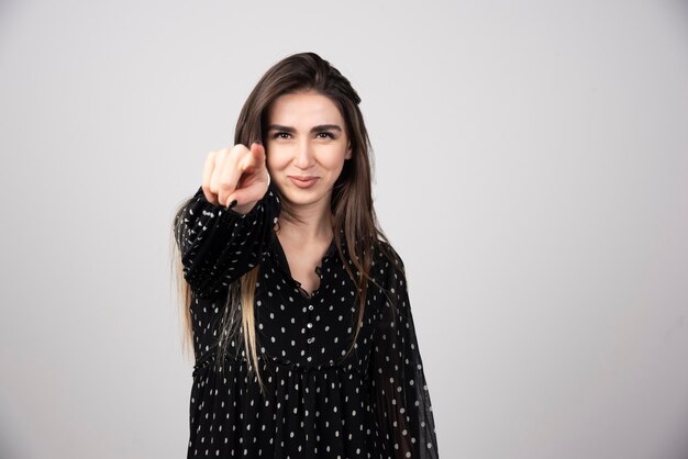 Glimlachende vrouw wijzend naar de camera op een grijze muur.