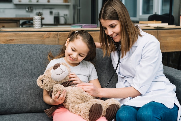 Glimlachende vrouwelijke arts die de teddybeergreep door gelukkig meisje onderzoekt