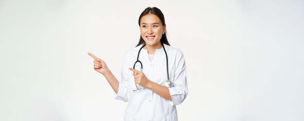 Glimlachende vrouwelijke Aziatische arts in medisch uniform wijzende vingers en kijkend naar links naar advertentie kopie ruimte promo staande in gewaad tegen witte achtergrond