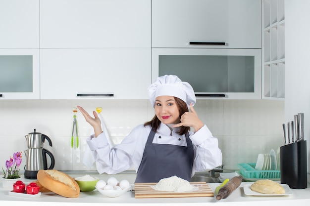Glimlachende vrouwelijke chef-kok in uniform die achter de tafel staat met snijplank broodgroenten die me een gebaar maken in de witte keuken