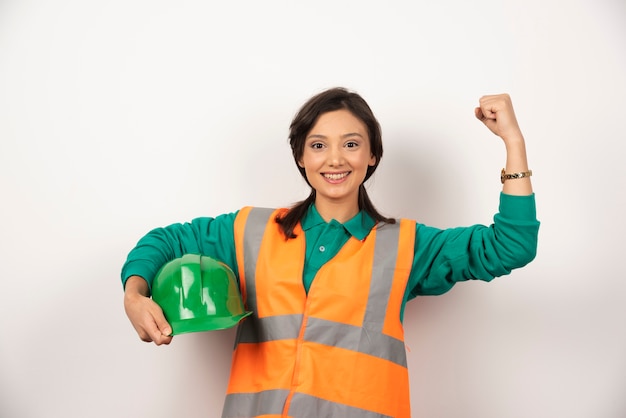 Glimlachende vrouwelijke ingenieur die spieren toont en een helm op witte achtergrond houdt