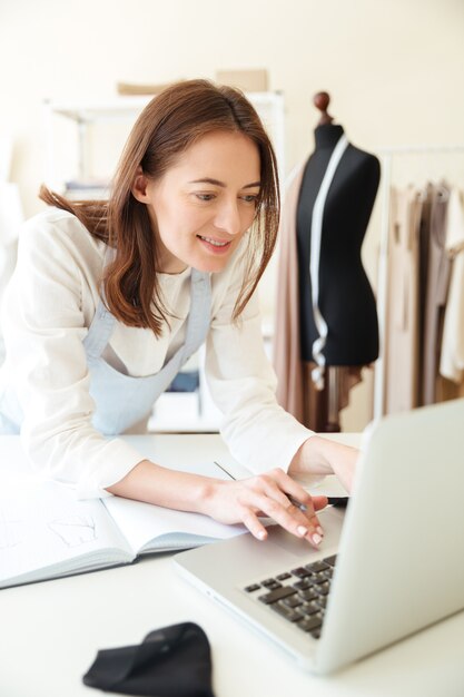 Glimlachende vrouwennaaister in het werken met laptop en stoffen in workshop