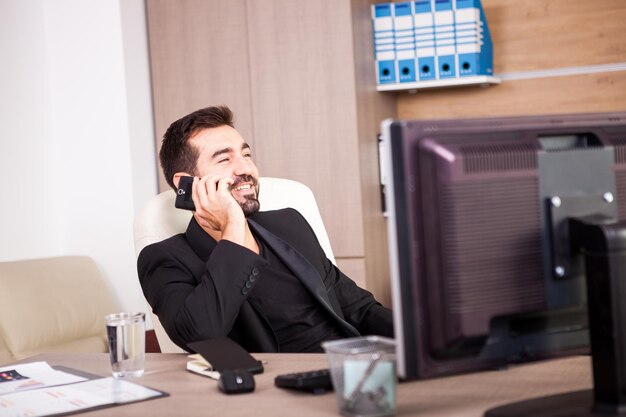 Glimlachende zakenman die aan de telefoon praat terwijl hij in zijn kantoor werkt. Ondernemer in professionele omgeving