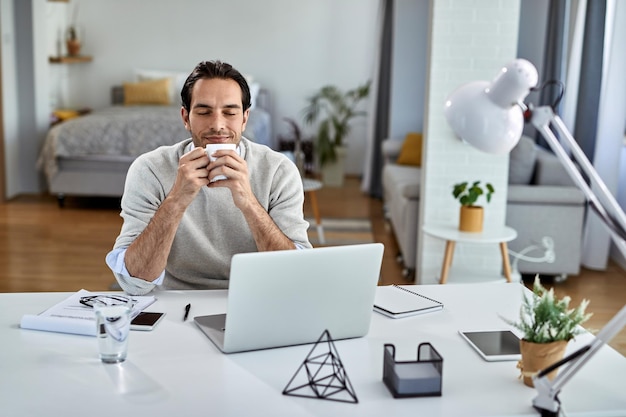 Gratis foto glimlachende zakenman die geniet van een koffiepauze met zijn ogen dicht terwijl hij thuis werkt