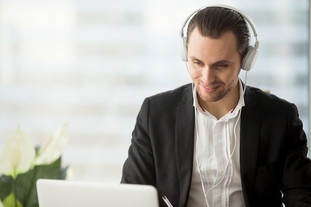Glimlachende zakenman die in hoofdtelefoon het laptop scherm bekijkt.