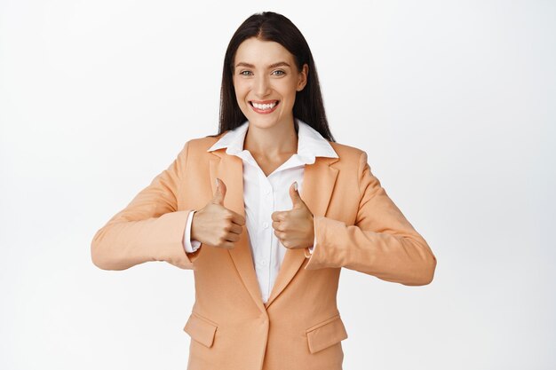Glimlachende zakenvrouw die duimen omhoog laat zien, keurt iets goeds goed Verkoopster geeft positieve feedback in pak tegen een witte achtergrond
