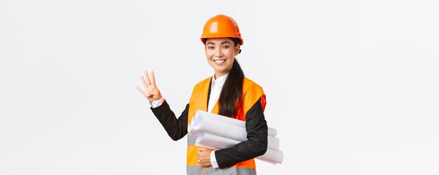 Glimlachende zelfverzekerde aziatische vrouwelijke ingenieur bouwmanager in veiligheidshelm draagt blauwdrukken showi