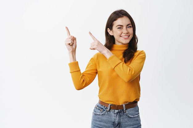 Glimlachende zelfverzekerde dame die advertentie op kopieerruimte toont en met de vingers naar de linkerbovenhoek wijst en er gelukkig uitziet om een speciale aanbieding te promoten die op een witte achtergrond staat