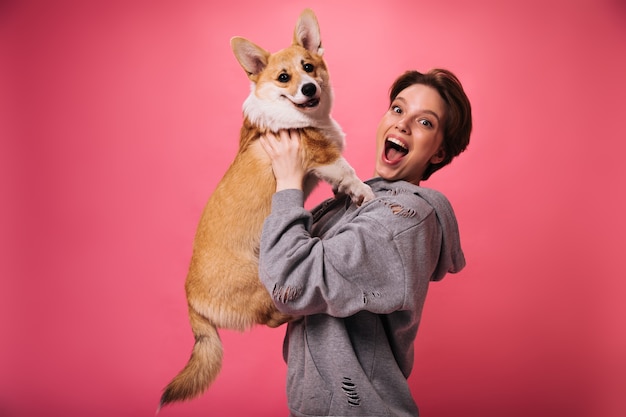 Goed gehumeurd vrouw houdt hond en lachen op roze achtergrond. Emotionele sort-haired meisje in grijze hoodie vormt met corgi op geïsoleerd