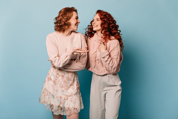 Goed geklede dames die op blauwe achtergrond praten. Studio die van aantrekkelijke meisjes is ontsproten die elkaar bekijken.