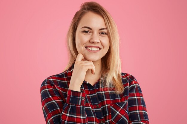 Goed uitziende blonde vrouw met zachte glimlach, houdt een hand onder de kin, gekleed in een geruit overhemd