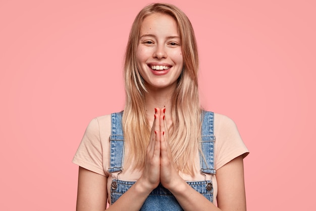 Gratis foto goed uitziende jonge langharige vrouw heeft een brede stralende glimlach, houdt de handpalmen tegen elkaar gedrukt, smeekt om iets in haar vriendje