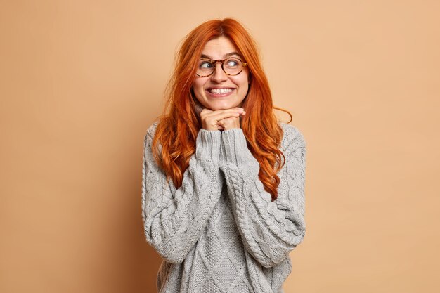 Goed uitziende jonge vrouw met rood haar houdt handen onder de kin glimlacht toothily geconcentreerd opzij draagt bril en trui.