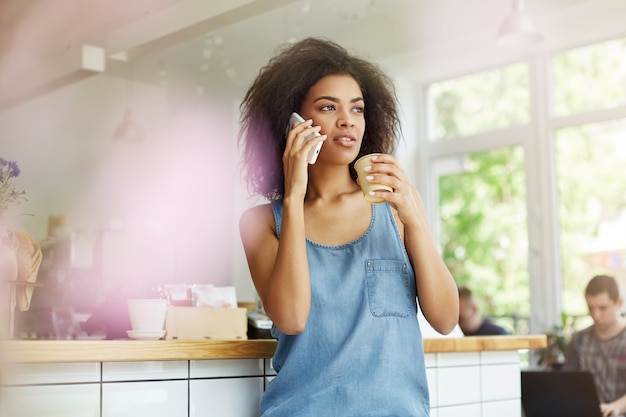 Goed uitziende jonge zwarte student Afrikaanse zwarte vrouw met krullend donker haar in casual stijlvolle kleding zittend in coffeeshop, espresso drinken, opzij kijken met bezorgde uitdrukking, praten over smartph