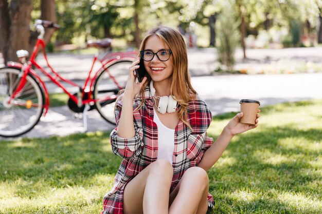 Goedgehumeurd Kaukasisch meisje dat vriend roept terwijl het drinken van koffie in park. Buitenfoto van geïnspireerde dame die op het gras rust.