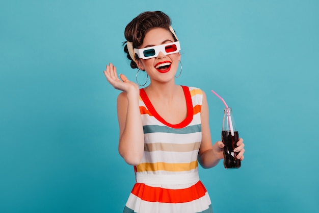 Goedgehumeurd pinupmeisje poseren in 3d-bril. studio shot van zalige brunette vrouw met fles frisdrank geïsoleerd op blauwe achtergrond.