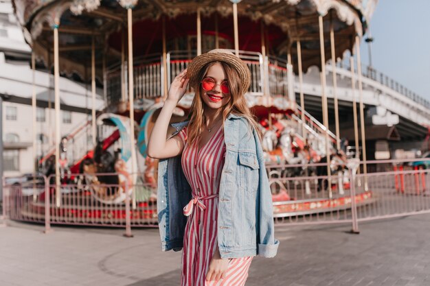 Goedgehumeurd vrouwelijk model in een strohoed die aan het koelen is in de buurt van de carrousel. Trendy zorgeloos meisje dag doorbrengen in pretpark.