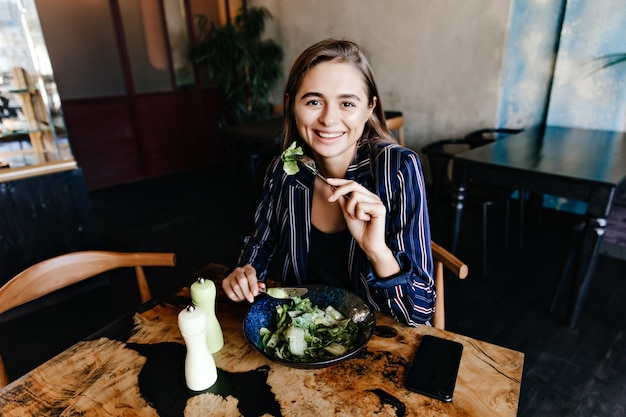 Gratis foto goedgehumeurde blanke vrouw die van gezond voedsel geniet. binnen schot van glimlachend meisje dat salade eet.