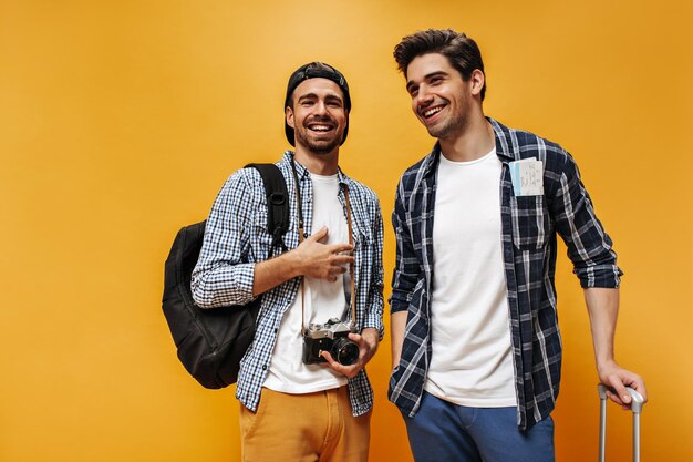 Goedgehumeurde jonge brunet mannen in geruite shirts en witte t-shirts glimlach op oranje achtergrond vrolijke reizigers poseren met retro camera en rugzak