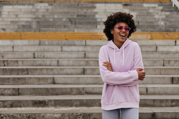 Goedgehumeurde jonge brunette krullend donkerhuidige vrouw in roze zonnebril en stijlvolle paarse hoodie glimlacht kruist armen en poses in de buurt van trappen