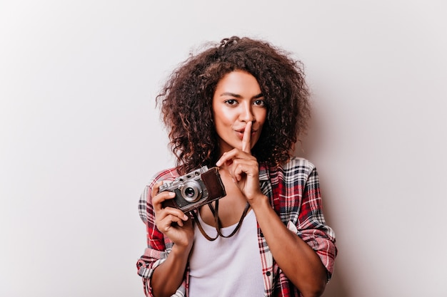 Goedgehumeurde jonge dame met camera die zich met glimlach bevindt. verfijnd zwart meisje dat foto's maakt.