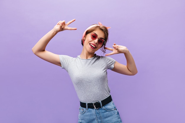 Goedgehumeurde vrouw in grijs overhemd en zonnebril die vredestekens tonen. Mooi meisje in roze bril en denim trendy rok poseren.