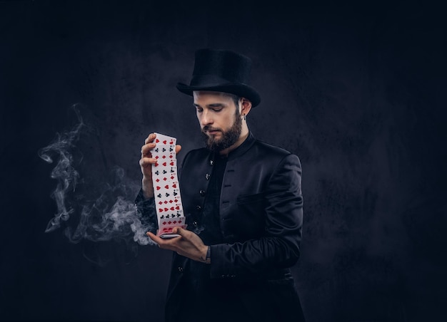 Goochelaar in een zwart pak en hoge hoed, met truc met speelkaarten en magische rook op een donkere achtergrond.