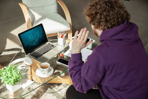 Gratis foto grafieken controleren. man aan het werk vanuit huis tijdens coronavirus of covid-19 quarantaine, extern kantoorconcept. jonge zakenman, manager die taken doet met smartphone, laptop, tablet heeft online conferentie.