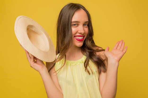 Grappig charmant meisje met roze lippen zwaait naar de camera terwijl ze een hoed vasthoudt en poseert voor de camera over een gele achtergrond