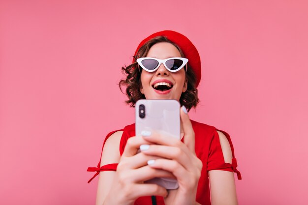 Grappig Kaukasisch meisje in Franse outfit met telefoon voor selfie. Lachende brunette dame in rode baret foto van zichzelf nemen.