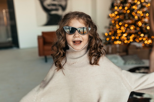 Grappig lief meisje met krullen dragen oversized gebreide trui en zonnebril dansen voor kerstboom