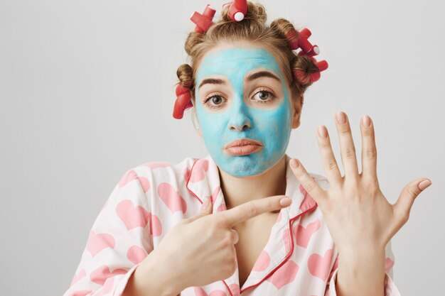 Grappig meisje in gezichtsmasker en haarkrulspelden wijzend op vinger zonder trouwring