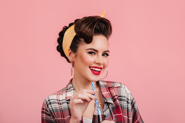 Gratis foto grappig pinupmeisje dat op roze achtergrond glimlacht. studio shot van trendy brunette vrouw in chckered shirt.